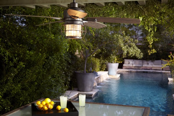 Pictured is an outdoor fan hanging from a pergola placed near a gorgeous pool with coastal architecture.